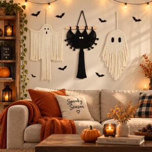 May include: Three macrame wall hangings featuring two off-white ghosts and one black bat, all suspended on a wall. The ghosts have simple black eyes, while the bat has two light-colored circular eyes and a long central tassel hanging downward.