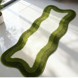 May include: A wavy, green and white rug with a unique shape. The rug features a central white section bordered by layers of varying green shades. The rug's edges are a darker green, creating a distinct border. The rug is placed on a white floor.