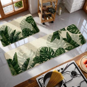 May include: A rectangular and a runner rug with a tropical leaf design. The rugs feature large green leaves on a beige background. The runner rug is long and narrow, while the other rug is rectangular.