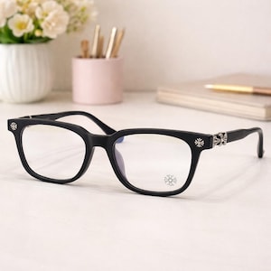 May include: Black rectangular-framed eyeglasses with silver-coloured cross-shaped detailing on the temples and lower right lens. The glasses are on a white surface with a blurred background.