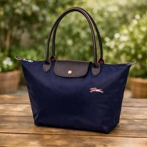 Puede incluir: Bolso tote azul marino con asas de cuero marrón y cierre de solapa. El bolso presenta un logotipo de caballo bordado en rosa y un cierre de cremallera. El bolso está sobre una superficie de madera.