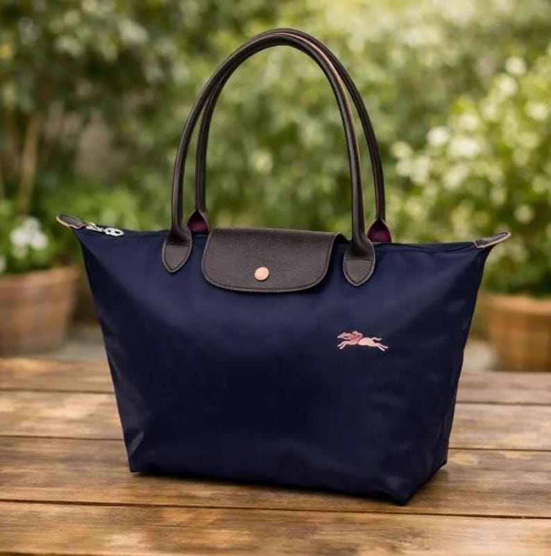 Puede incluir: Bolso tote azul marino con asas de cuero marr&oacute;n y cierre de solapa. El bolso presenta un logotipo de caballo bordado en rosa y un cierre de cremallera. El bolso est&aacute; sobre una superficie de madera.