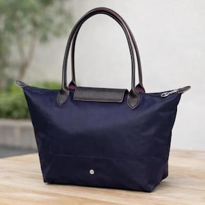Puede incluir: Bolso tote de color azul marino con asas de cuero negro y solapa marr&oacute;n oscuro. El bolso tiene una cremallera en la parte superior y un peque&ntilde;o bot&oacute;n plateado en la parte delantera. Las asas son arqueadas y est&aacute;n unidas al bolso con detalles de cuero marr&oacute;n oscuro.