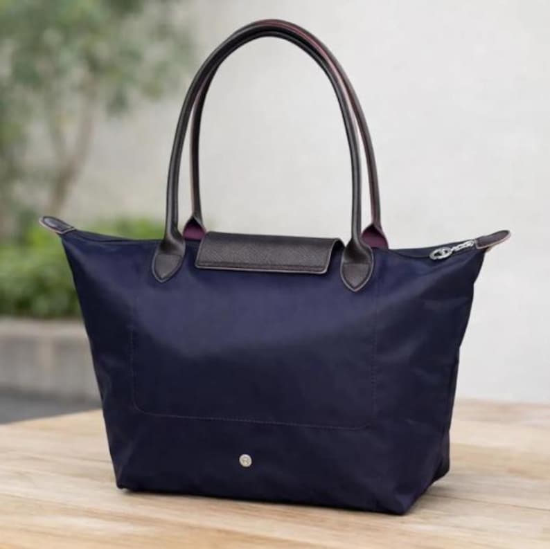 Puede incluir: Bolso tote de color azul marino con asas de cuero negro y solapa marr&oacute;n oscuro. El bolso tiene una cremallera en la parte superior y un peque&ntilde;o bot&oacute;n plateado en la parte delantera. Las asas son arqueadas y est&aacute;n unidas al bolso con detalles de cuero marr&oacute;n oscuro.