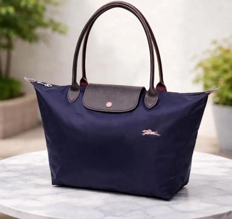 Puede incluir: Bolso tote azul marino con asas de cuero marr&oacute;n y cierre de solapa. El bolso presenta un logotipo rosa de un caballo corriendo y un bot&oacute;n rosa en la solapa. El bolso es de nailon.