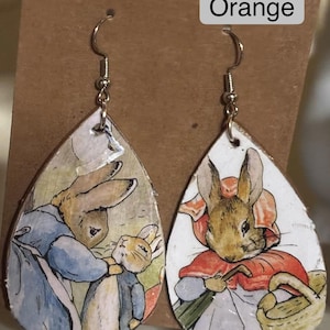 Può includere: Orecchini a goccia con illustrazioni di conigli in stile libro di fiabe. Gli orecchini hanno ganci color argento e presentano scene di conigli vestiti. Il testo "5 Orange" è visibile in alto a destra.