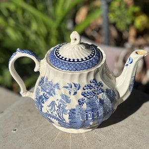 Puede incluir: Tetera de cerámica vintage con un diseño azul y blanco de patrón Willow. La tetera presenta un asa curva, un pico y una tapa con un adorno decorativo. El diseño incluye escenas de pagodas, árboles y pájaros.