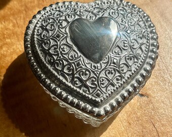 May include: A silver heart-shaped trinket box with ornate filigree detailing and a smooth heart in the center. The box has a beaded border and sits on a wooden surface. The box is likely used for storing small jewelry items.