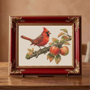 May include: Cross-stitch artwork of a red cardinal perched on a branch with apples. The framed piece has a red frame with gold accents. The artwork is displayed on a wooden surface.