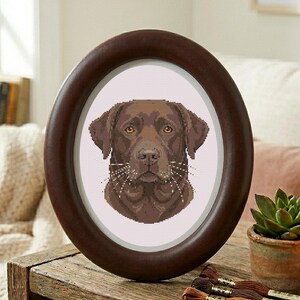 May include: A framed cross-stitch portrait of a chocolate Labrador Retriever dog. The oval frame is dark brown, and the dog's image is centered on a white background. The dog's fur is brown, with detailed stitching.
