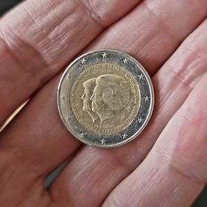 May include: A two euro coin with a gold centre and a silver outer ring. The coin shows a profile of two figures and the text "KONING DER NEDERLANDEN" and "2012". The coin is held in a hand.