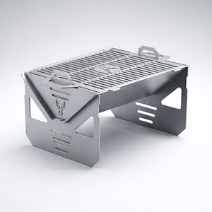 May include: A silver-coloured, portable folding grill with a rectangular cooking surface and a deer head logo. The grill has a slotted grate, handles, and cut-out designs on the sides. It is designed for outdoor cooking and camping.