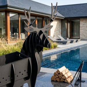 May include: A matte black metal charcoal grill shaped like a deer with large antlers. The grill features a rectangular firebox with perforated side panels for ventilation and a sturdy base. A stack of firewood and metal tongs are placed nearby.
