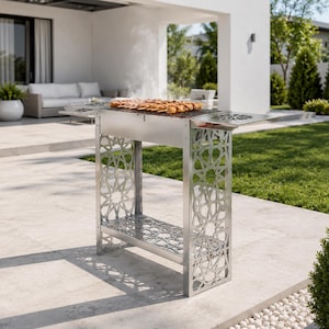 Può includere: Un barbecue da esterno in acciaio inossidabile con un motivo geometrico decorativo sui lati e un ripiano inferiore. Il barbecue rettangolare ha una superficie di cottura e un lato incernierato. Spiedini di cibo vengono cotti sulla griglia, con fumo che sale.
