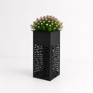 May include: A tall, square, matte black metal planter featuring a laser-cut pattern of horizontal wavy lines on its four sides. The planter is filled with a dense cluster of green foliage and small, light pink flowers blooming at the top.