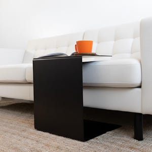 May include: A black, C-shaped side table stands beside a white sofa. An open book, a small wooden coaster, and an orange mug are on the table's surface. The table is made of metal and has a minimalist design. The table's height is approximately 60 cm.