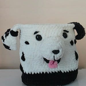 May include: A white and black crocheted storage basket shaped like a dalmatian dog. The basket features a protruding snout, a small pink tongue, black plastic eyes, and floppy ears with black spots. The base of the basket is solid black.
