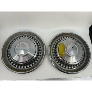 May include: Two circular chrome Chevrolet Motor Division hubcaps featuring a central emblem and a ring of decorative cutouts. The metallic surfaces have a polished finish with a distinct circular design pattern radiating from the center.