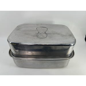 May include: A rectangular silver-colored metal roasting pan with a matching lid. The lid features a central wire loop handle for lifting. The pan has rounded corners and a smooth, polished finish, showing a two-piece construction with a base and lid.