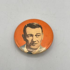 May include: Circular orange button with a portrait of a man in a suit. The man's face is centrally positioned, with his hair neatly styled. The button has a simple design and a vintage aesthetic.