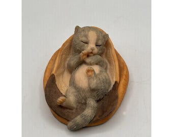 May include: A figurine of a sleeping gray and white kitten curled up in a brown and tan basket. The kitten is lying on its back with its eyes closed and paws near its face. The basket has a curved shape.