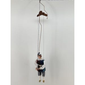 May include: A vintage marionette puppet with a painted face, wearing a white shirt, blue and burgundy vest, and grey trousers. The puppet is suspended by strings from a wooden control bar.