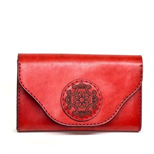 May include: A red leather wallet with a flap closure. The wallet features a circular, embossed design in the center and black stitching around the edges. The wallet measures approximately 7 inches long.