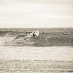 May include: Sepia-toned image of surfers riding waves. One surfer is mid-air, performing a trick on a breaking wave. Other surfers are visible in the background, enjoying the surf. The sea is choppy.
