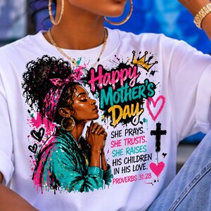 May include: A white t-shirt featuring a graphic illustration of a person with a headscarf and curly hair. The text reads Happy Mother's Day, She Prays, She Trusts, She Raises His Children In His Love, Proverbs 31:28, with hearts and a cross.