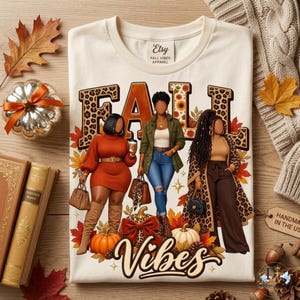 May include: A cream-colored t-shirt featuring a graphic of three individuals and the text FALL VIBES. The design includes leopard print lettering, autumn leaves, pumpkins, and stylized clothing. The shirt has a tag reading Etsy FALL VIBES APPAREL.