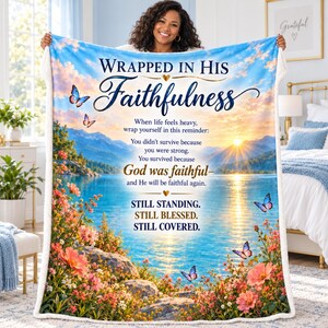 May include: A rectangular fleece blanket featuring a scenic lake and mountain landscape at sunrise with colorful butterflies and floral borders. The text reads: WRAPPED IN HIS Faithfulness, When life feels heavy, wrap yourself in this reminder: You didn't survive because you were strong. You survived because GOD was faithful—and He will be faithful again. STILL STANDING. STILL BLESSED. STILL COVERED.
