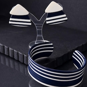 May include: A navy blue and white striped cuff bracelet and matching earrings. The earrings are triangular with white tops. The jewellery is displayed on a clear stand and a dark, textured surface. The bracelet is open.