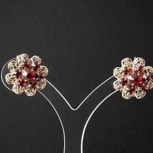 May include: A pair of vintage-style earrings. Each earring features a gold-toned filigree design with a central, large, faceted, red gemstone surrounded by smaller red stones. The earrings are displayed on a white stand against a black background.