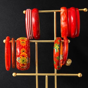 Peut inclure: Un présentoir à bracelets en métal doré contenant divers bracelets rouges et oranges. Certains bracelets présentent des motifs floraux, des rayures ou des motifs marbrés. Le fond est noir uni.