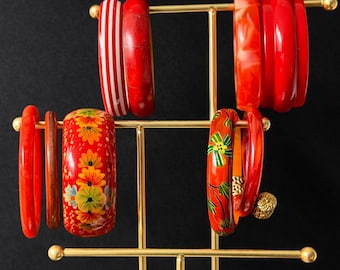 May include: A gold-colored metal bracelet display stand holding various red and orange bangles. Some bangles have floral patterns, stripes, or marbled designs. The background is a solid black.