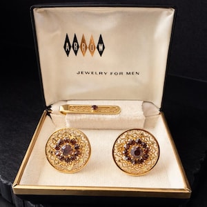 May include: Vintage gold-toned jewelry set in a black and gold box. The set includes a tie clip and a pair of cufflinks, each featuring a central, round, dark red stone surrounded by an ornate, openwork design. The box is labeled "ARROW JEWELRY FOR MEN."