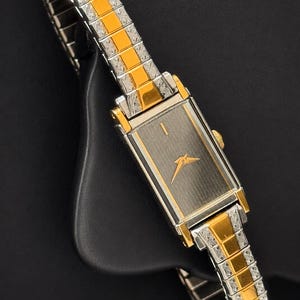 May include: A rectangular wristwatch with a silver and gold-tone metal band and case. The watch face is dark gray with gold-tone hands and a small bird-like emblem. The band features a repeating pattern of gold and silver links.