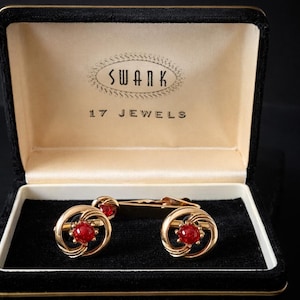 May include: Vintage gold-tone cufflinks with red gemstone centers, displayed in a black velvet-lined box. The box lid is open, revealing the "SWANK 17 JEWELS" logo. The cufflinks have a circular design with intricate detailing.
