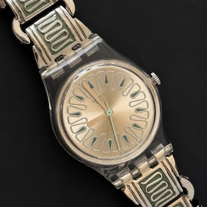 May include: A Swatch watch with a silver-tone metal band and a clear plastic case. The watch face is gold with green accents and a green second hand. The band features a repeating green and silver design.