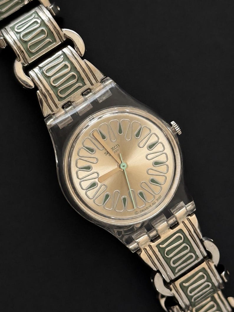 May include: A Swatch watch with a silver-tone metal band and a clear plastic case. The watch face is gold with green accents and a green second hand. The band features a repeating green and silver design.