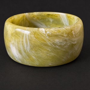 May include: A wide bangle bracelet featuring a polished, smooth surface with a swirling marbled pattern in shades of olive green, chartreuse, and bright white. The circular accessory has a thick, rounded profile and a glossy, reflective finish.