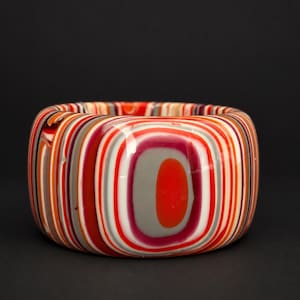 May include: A thick, cylindrical bangle bracelet featuring a multi-layered, concentric pattern of vibrant orange, deep red, white, and soft grey stripes. The surface is smooth and glossy, displaying a repeating, rounded rectangular design.