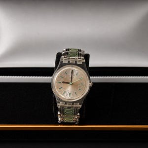 May include: A silver-toned wristwatch with a light-colored face and green accents on the band, displayed in a black presentation box with a white interior. The watch features a clear band and a light-colored face with green hands.