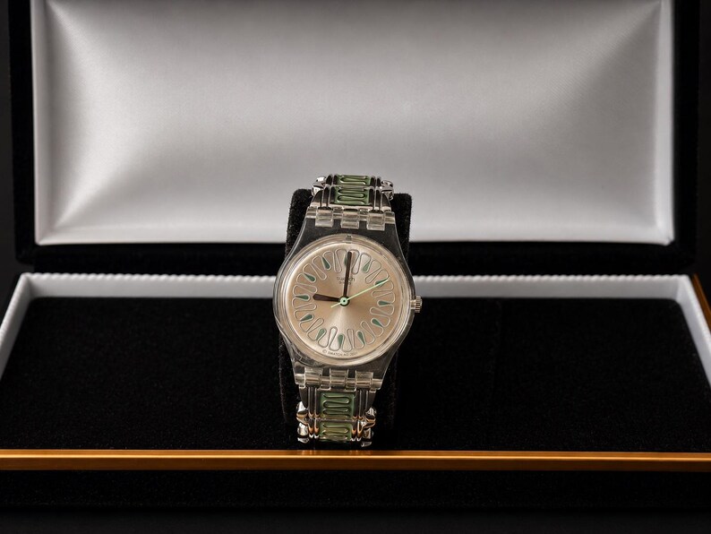 May include: A silver-toned wristwatch with a light-colored face and green accents on the band, displayed in a black presentation box with a white interior. The watch features a clear band and a light-colored face with green hands.