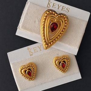 May include: A gold-tone brooch and matching pair of clip-on earrings, each shaped as a heart with a beaded border and a central red teardrop-shaped stone. The jewelry is displayed on white cards labeled with the brand name KEYES in gold lettering.