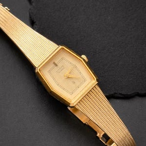 May include: A gold-toned Citizen Quartz watch with a rectangular face and a matching mesh band. The watch face has a cream-colored background with gold hands and markers. The watch is displayed on a dark surface.