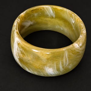 May include: A wide, circular bangle bracelet featuring a polished, translucent finish with a marbled pattern of olive green, mustard yellow, and streaks of bright white throughout the entire surface of the thick, rounded resin material.