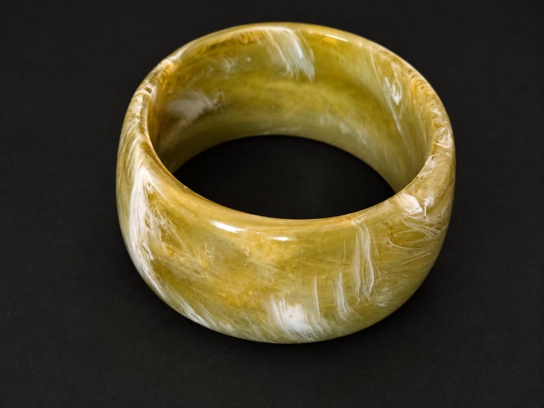 May include: A wide, circular bangle bracelet featuring a polished, translucent finish with a marbled pattern of olive green, mustard yellow, and streaks of bright white throughout the entire surface of the thick, rounded resin material.