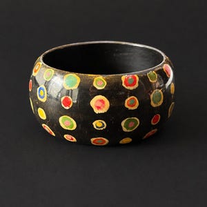 May include: A wide, dark brown bangle bracelet featuring a repeating pattern of hand-painted, multi-colored circular dots in shades of red, yellow, green, and blue, each outlined with a thin, light-colored border against a dark, smooth surface.
