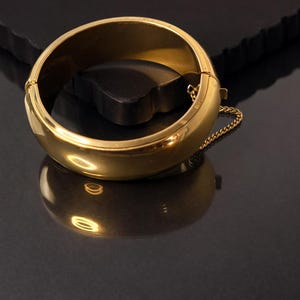 May include: A gold-coloured hinged bangle bracelet with a safety chain. The bracelet is round and has a smooth, polished surface. The bracelet is resting on a dark surface, reflecting the light.